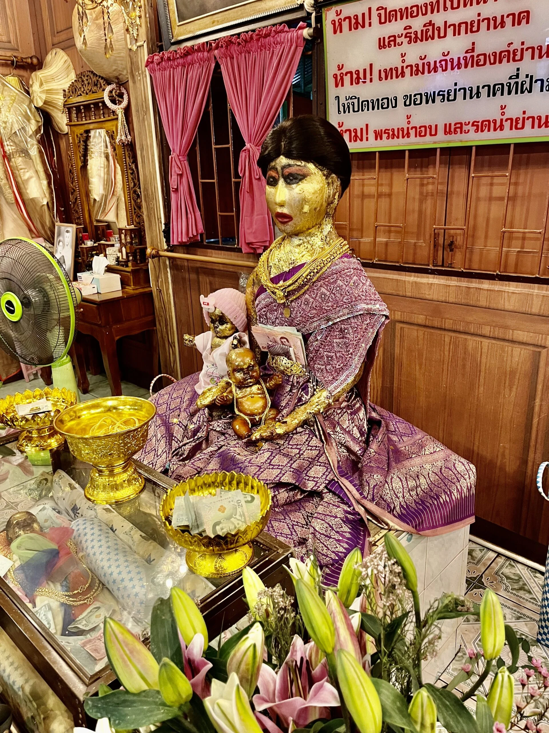 A statue of a women in Thai costume holding a baby, both are covered in gold leaf. The woman is sitting on a platform in a wooden house. There are gold offering trays on a table in front of her, as well as flowers.