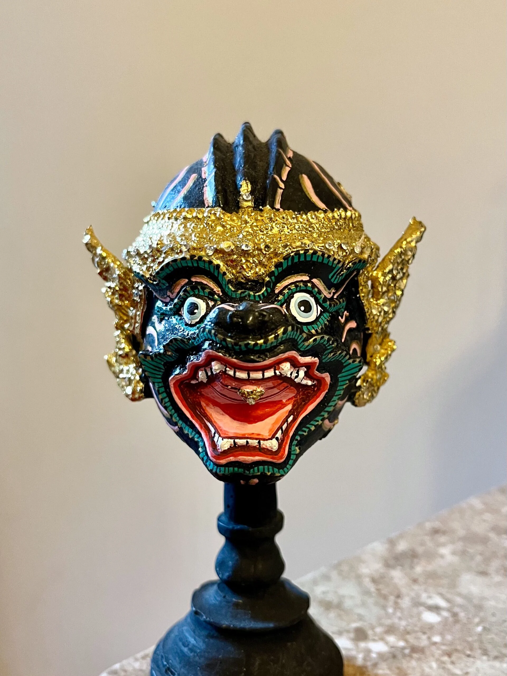 A black mask of Hanuman with a golden headdress with an open mouth and pointed teeth.