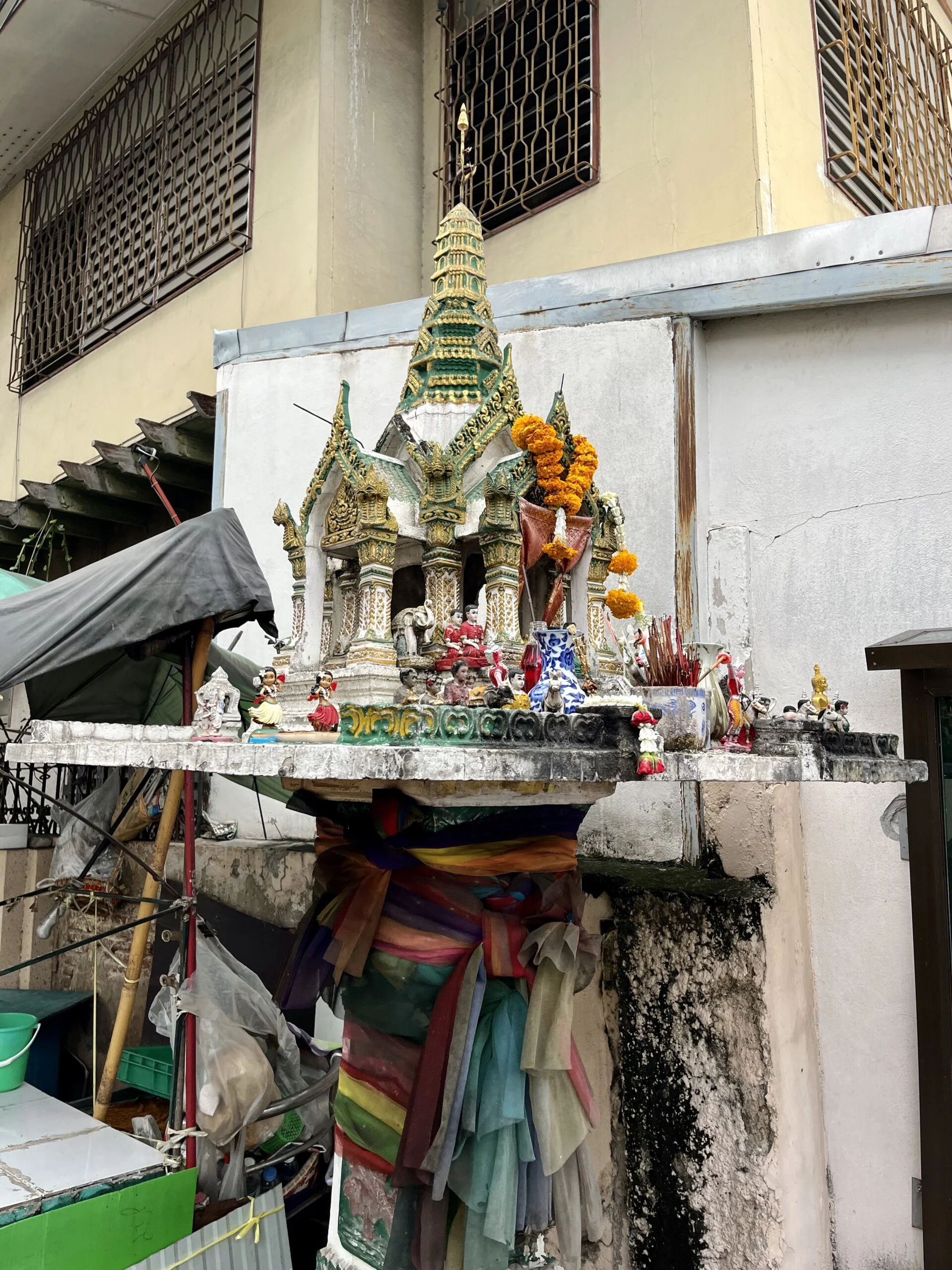 A white shrine resembling a temple structure with a pointed roof sits on a single pillar. Old pieces of coloured cloth are wrapped around the pillar.