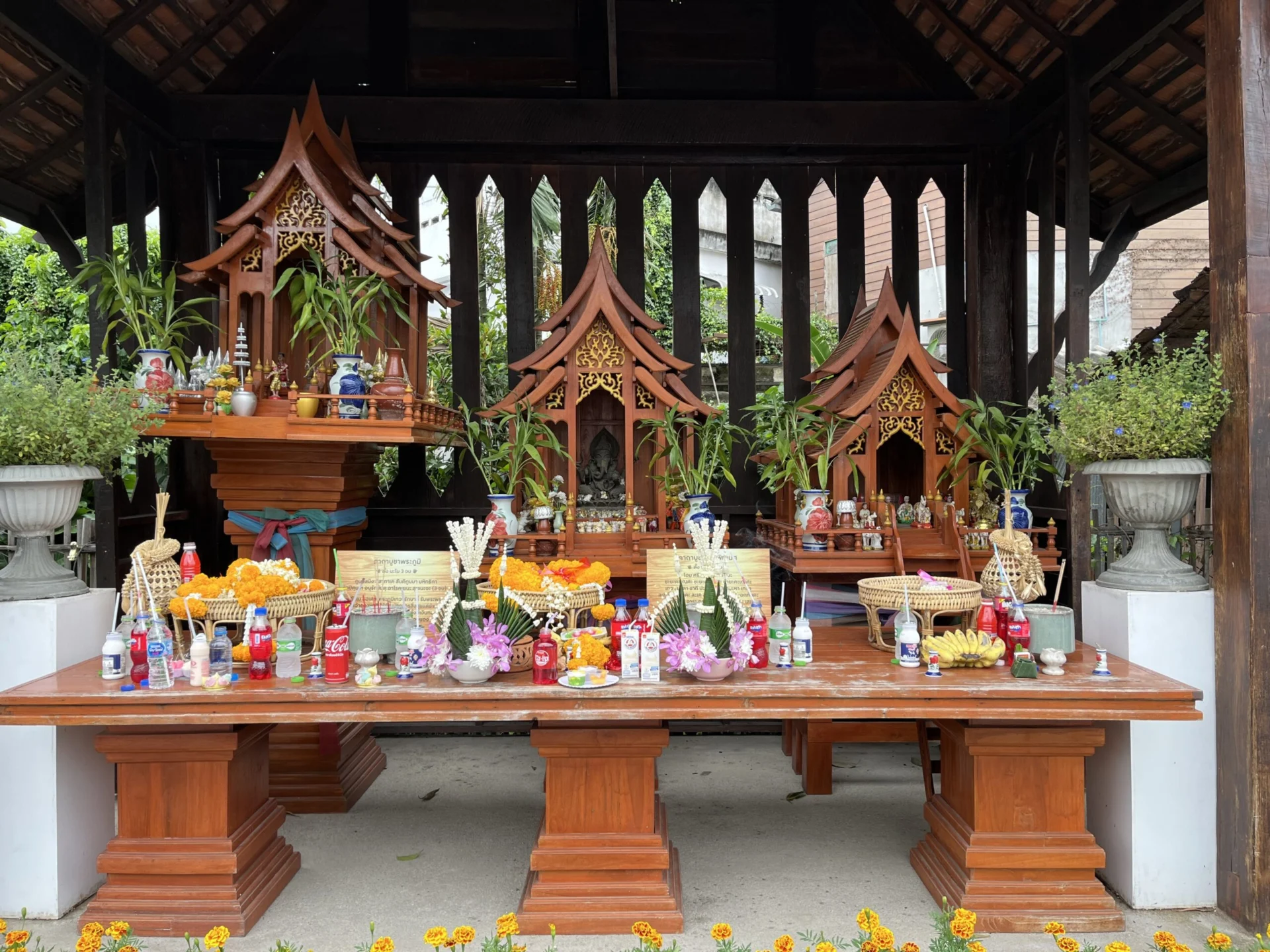 Three wooden shrines in a row, from largest on the left to smallest on the right. Each has offerings of flowers, food, and drink in front of them. They each resemble a traditional Thai-style house or temple, with a triangular, pointed roof.