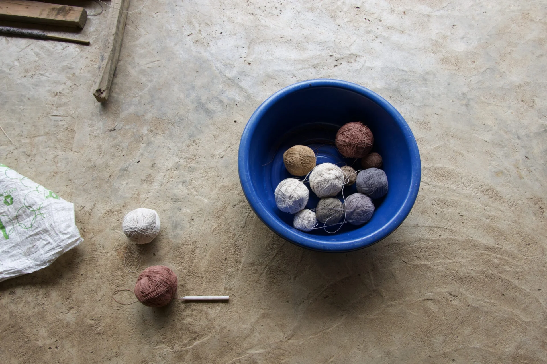 A bright blue bucket is placed on a concrete floor. Two spools of thread are on the ground next to it – one white, one brown. In the basket are more spools of various colours.