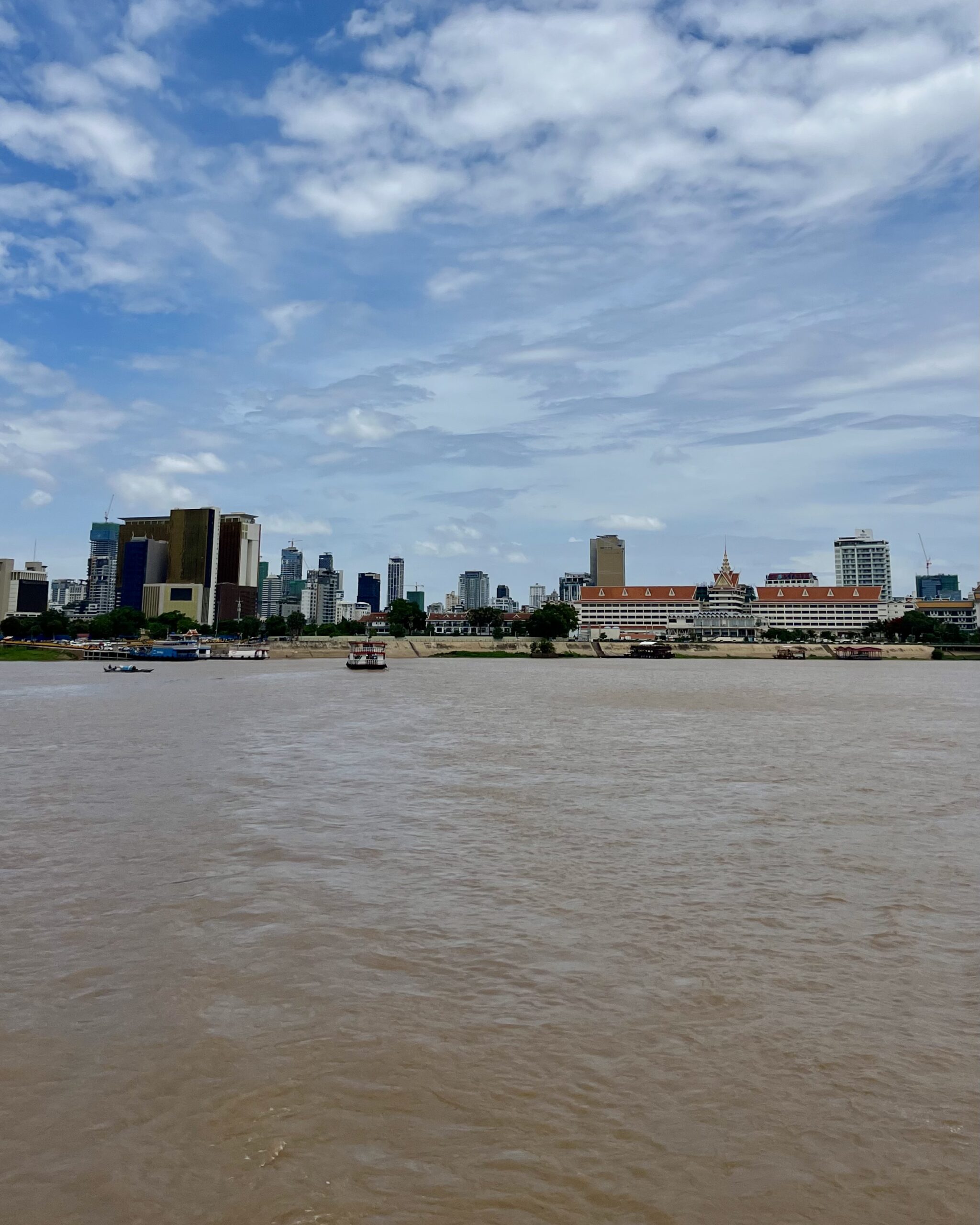 A vast brown body of water with a cityscape in the background and a bright blue sky wit white clouds.