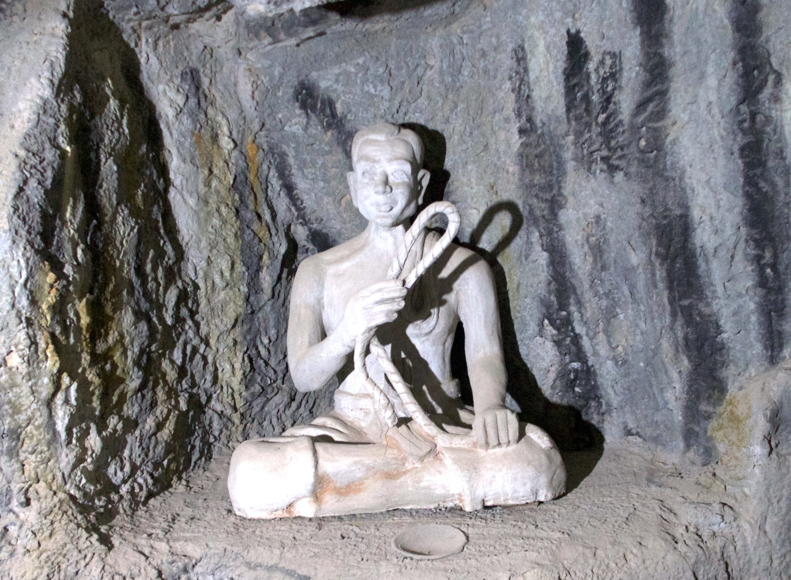 Statue of a man sitting cross-legged, carved in white, holding up a lasso in his right hand.