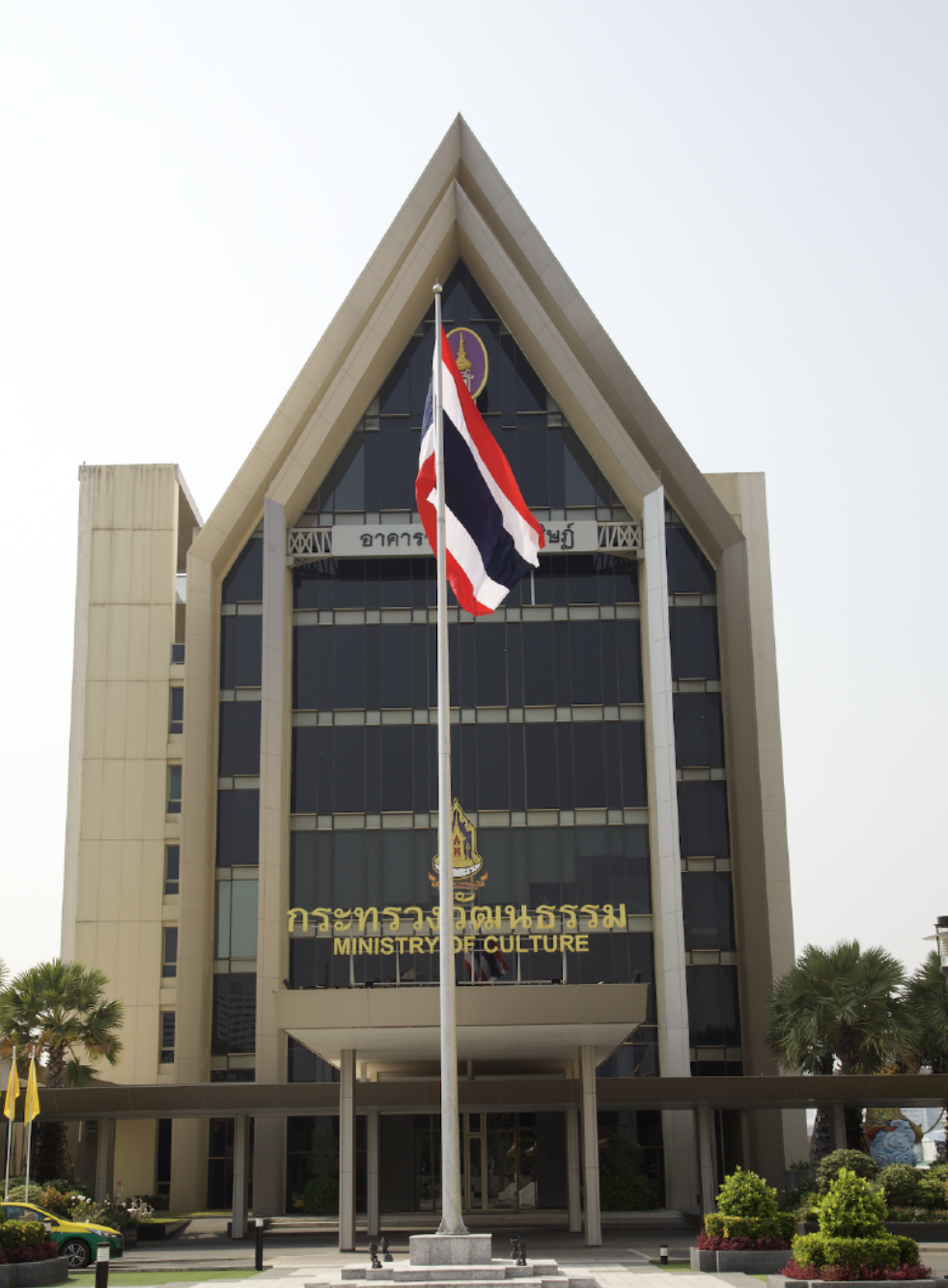 A building with large windows and a triangular roof. The sign reads: Ministry of Culture. There is a Thai flag with red, white, blue, white and red horizontal stripes on a flag pole in front of the building.
