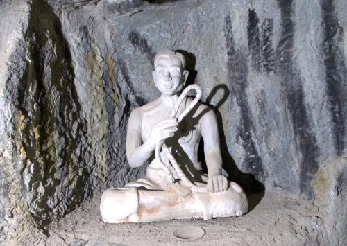 Statue of a man sitting cross-legged, carved in white, holding up a lasso in his right hand.