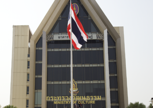 A building with large windows and a triangular roof. The sign reads: Ministry of Culture. There is a Thai flag with red, white, blue, white and red horizontal stripes on a flag pole in front of the building.